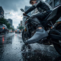 A person on a motorcycle in the rain wears a dark gray silicone shoe cover with a textured sole, splashing through a puddle.