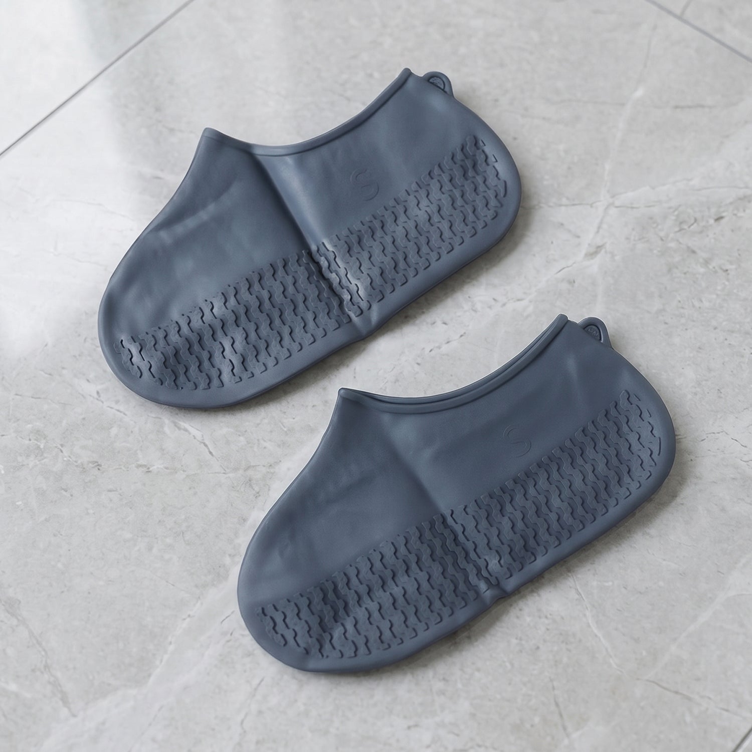 Two dark grey silicone shoe covers are laid flat on a light grey marble surface, showcasing their textured bottoms.